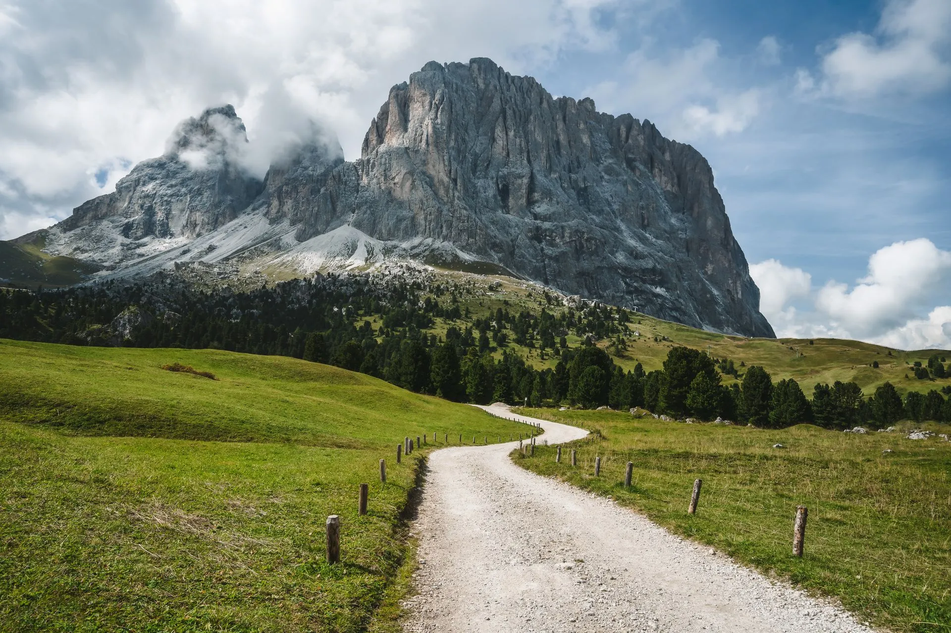 Schotterweg über grüne Almwiesen der Seiser Alm auf die Felswände von Langkofel und Plattkofel zu
